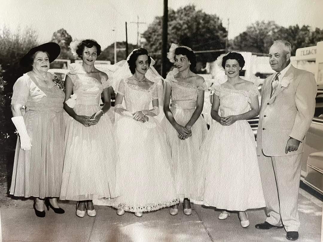 A wedding day in the 1950s — the bride in her veil flanked by three attendants in matching gowns, an older woman in a wide-brimmed hat at one end and a man in a pale suit at the other. Aunt Mabel is among them.