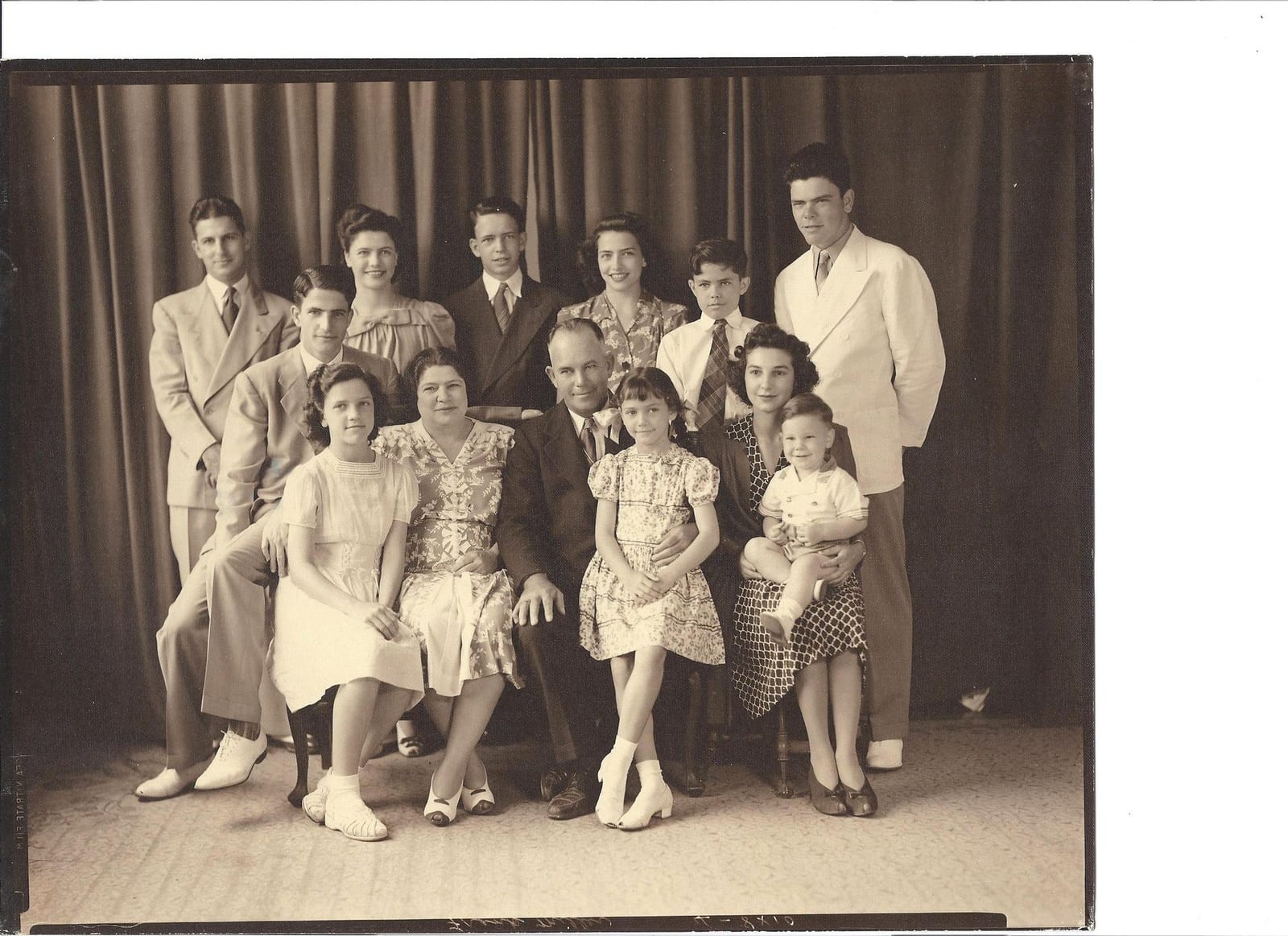 A formal sepia family portrait in front of a studio curtain — three generations in suits and patterned dresses, the grandparents seated at the centre with the youngest children in front. Aunt Mabel stands at the top left.